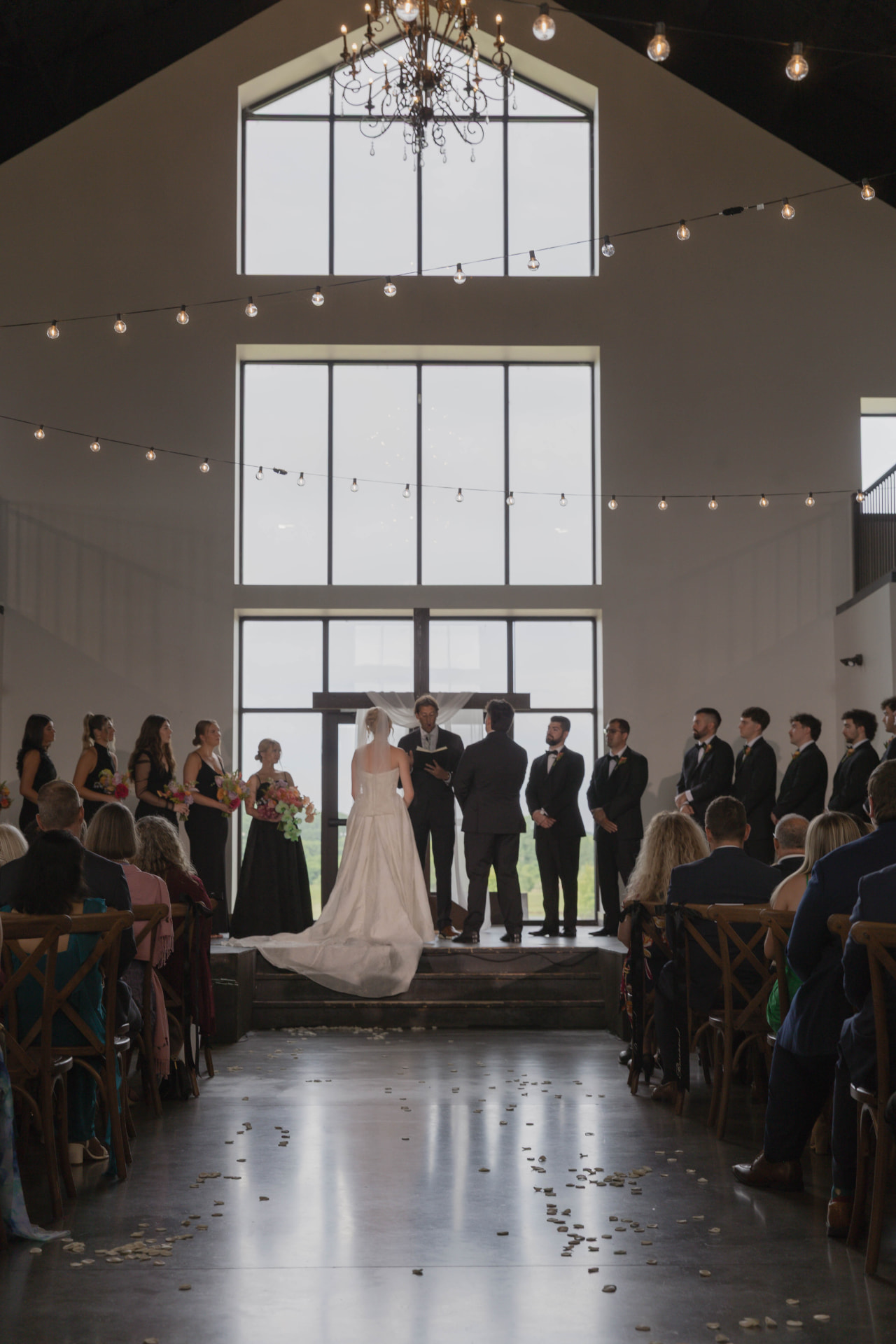 Couple exchanging vows beneath a chandelier and tall barn windows at a Columbia MO wedding venue